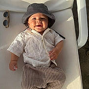Vincent is registered to the contest to win money with this photo: baby, child, smiling, hat, bucket_hat, shirt, embroidery, striped_pants, footwear, sunglasses, chair, white_chair, outdoor, sunlight, happy, portrait, infant, casual, relaxing, cute