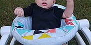 Winifred is registered to the contest to win money with this photo: baby, child, infant, bow, headband, black_shirt, baby_seat, colorful_pattern, grass, outdoor, legs, arms, fists, determined_expression, fisher_price, person, cute, young, sitting, playful
