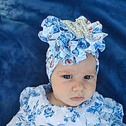 Winifred is registered to the contest to win money with this photo: baby, child, face, headwrap, clothing, blue, white, floral_pattern, portrait, serious_expression, infant, fabric, ruffles, cute, person, closeup, indoor, soft_background, headwear, eyebrows