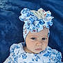 baby, child, face, headwrap, clothing, blue, white, floral_pattern, portrait, serious_expression, infant, fabric, ruffles, cute, person, closeup, indoor, soft_background, headwear, eyebrows