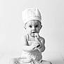 baby, chef_hat, apron, wooden_spoon, glass_jar, sitting, curious, monochrome, black_and_white, infant, cute, studio, portrait, child, kitchen, cooking, food, expression, smiling, playful