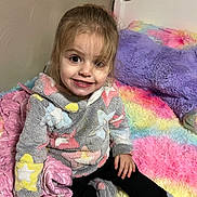 Lydia is registered to the contest to win money with this photo: child, girl, smiling, bed, colorful, fuzzy, hoodie, star_pattern, socks, indoor, happy, cute, portrait, young_child, plush, cross_legged, cozy, bedroom, soft_texture, casual_clothing