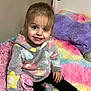 child, girl, smiling, bed, colorful, fuzzy, hoodie, star_pattern, socks, indoor, happy, cute, portrait, young_child, plush, cross_legged, cozy, bedroom, soft_texture, casual_clothing