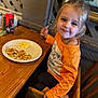 child, girl, smile, orange_shirt, high_chair, fork, plate, scrambled_eggs, toast, table, wooden_table, sippy_cup, restaurant, indoor, food, happy, meal, person, seated, wall_art