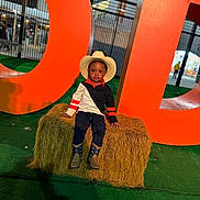 Tymair joined the competition — help win amazing prizes! child, boy, cowboy_hat, boots, hay_bale, orange_letters, outdoor, green_turf, night, portrait, casual_clothing, jeans, long_sleeve_shirt, fence, building, shadow, event, posing, serious_expression, urban