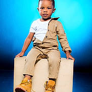 Tymair is registered to the contest to win money with this photo: child, toddler, sitting, boots, beige_clothing, white_tshirt, studio, blue_background, wooden_block, fashion, portrait, cute, young_child, confident, footwear, casual_wear, studio_lighting, expression, indoor, pose