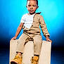 child, toddler, sitting, boots, beige_clothing, white_tshirt, studio, blue_background, wooden_block, fashion, portrait, cute, young_child, confident, footwear, casual_wear, studio_lighting, expression, indoor, pose