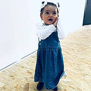 Lilia participe au concours pour gagner de l'argent avec cette photo : toddler, child, girl, denim_dress, white_bows, curly_hair, black_boots, wooden_floor, indoor, standing, hands_on_face, expression, cute, fashion, portrait, young_child, minimalist, wall, flooring, casual
