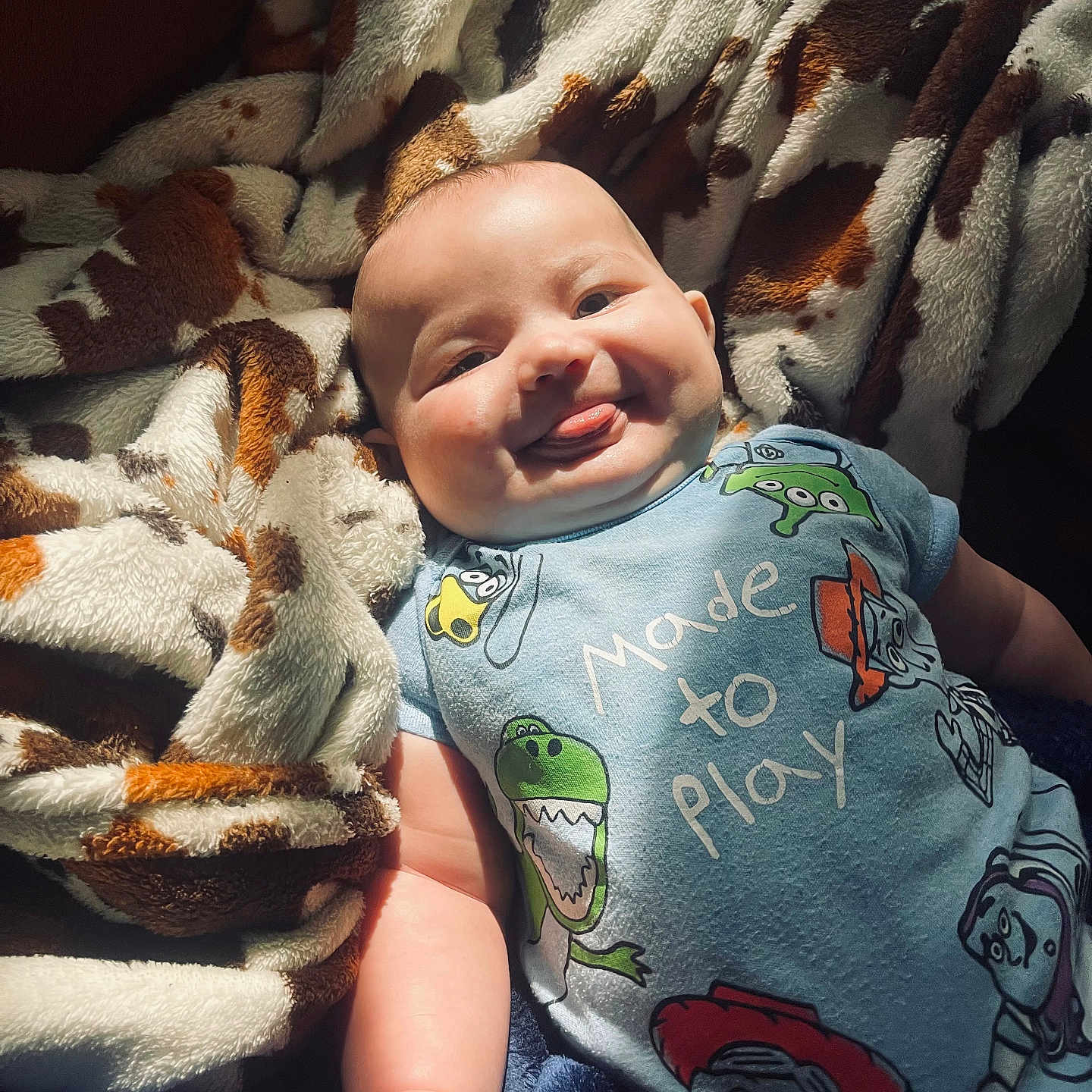 Kolsen is registered to the contest to win money with this photo: baby, blanket, cartoon_characters, child, chubby_cheeks, cozy, cute, happy, indoors, infant, light_and_shadow, lying_down, onesie, patterned_blanket, person, playful, portrait, smiling, soft_texture, tongue_out