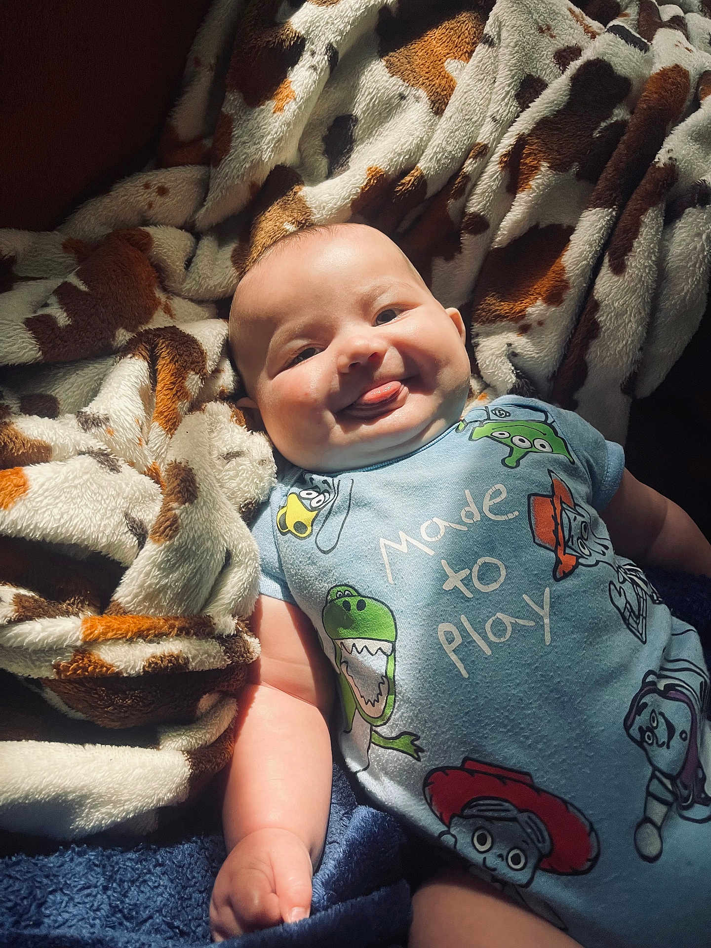 Kolsen is registered to the contest to win money with this photo: baby, infant, tongue_out, onesie, cartoon_characters, blanket, patterned_blanket, soft_texture, smiling, chubby_cheeks, cute, lying_down, playful, indoors, child, happy, portrait, light_and_shadow, cozy, person