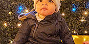 Oscar a rejoint le concours — aidez-le/la à gagner de superbes lots ! bear_ears, child, cozy, curious, cute, face, fairy_lights, golden_surface, greenery, hat, indoors, lights, portrait, puffy_jacket, sitting, snowsuit, toddler, warm_lighting, winter, winter_clothing