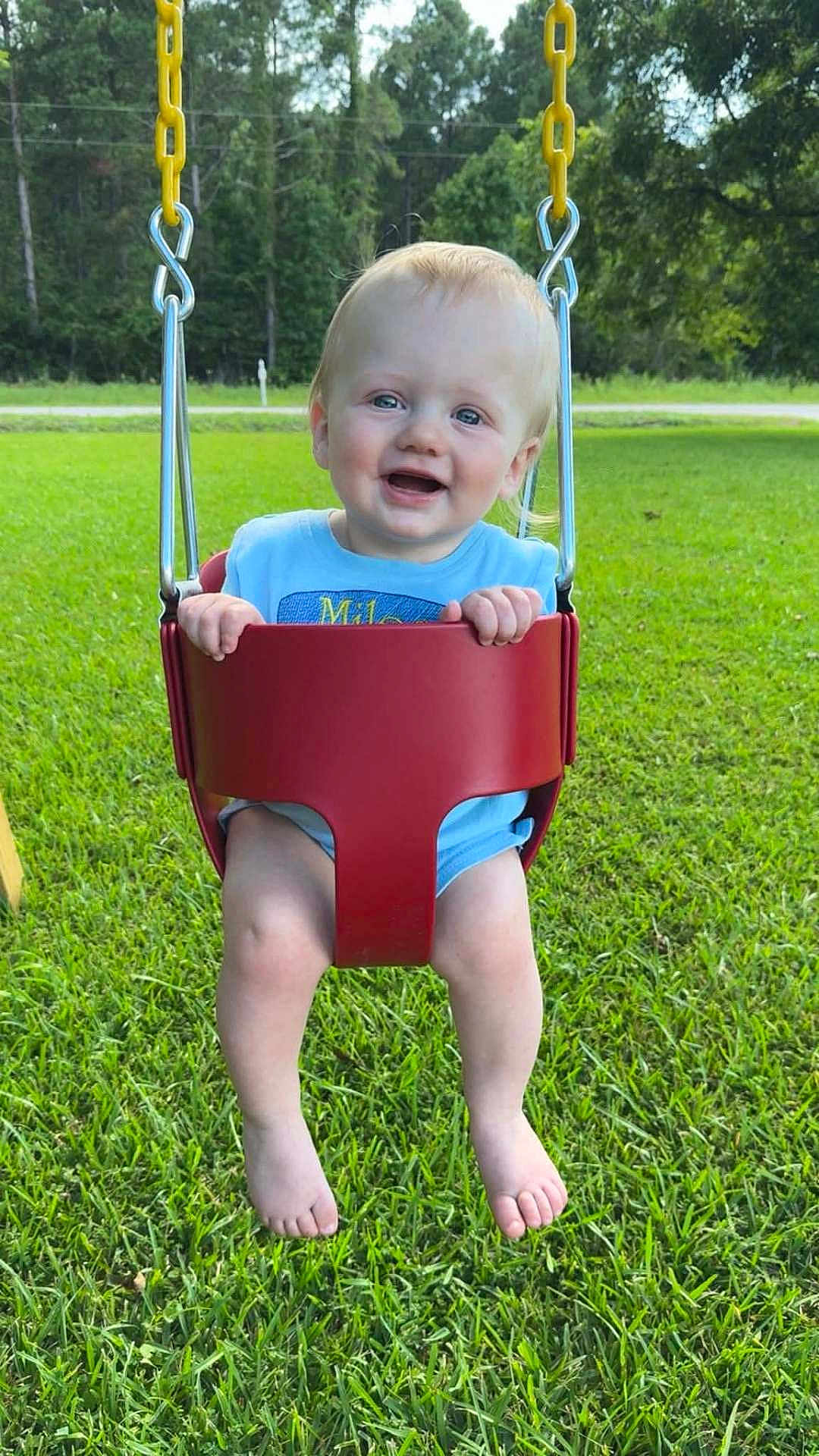 Miles is registered to the contest to win money with this photo: baby, child, swing, outdoor, grass, greenery, happy, smiling, blue_eyes, toddler, playground, sunny, nature, person, legs, barefoot, yellow_chain, red_swing, casual_clothing, daytime
