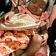Milan is registered to the contest to win money with this photo: baby, blanket, bottle, car_seat, child, close_up, cozy, curly_hair, dark_skin, heart_pattern, indoors, infant, peaceful, pink_clothing, portrait, rainbow_pattern, resting, seated, sleeping, textile