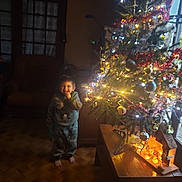 Claude a rejoint le concours — aidez-le/la à gagner de superbes lots ! barefoot, chair, child, christmas_tree, cozy, decorations, festive, furniture, holiday, indoors, lamp, lights, ornaments, pajamas, person, smile, smiling, warm_lighting, window, wooden_floor
