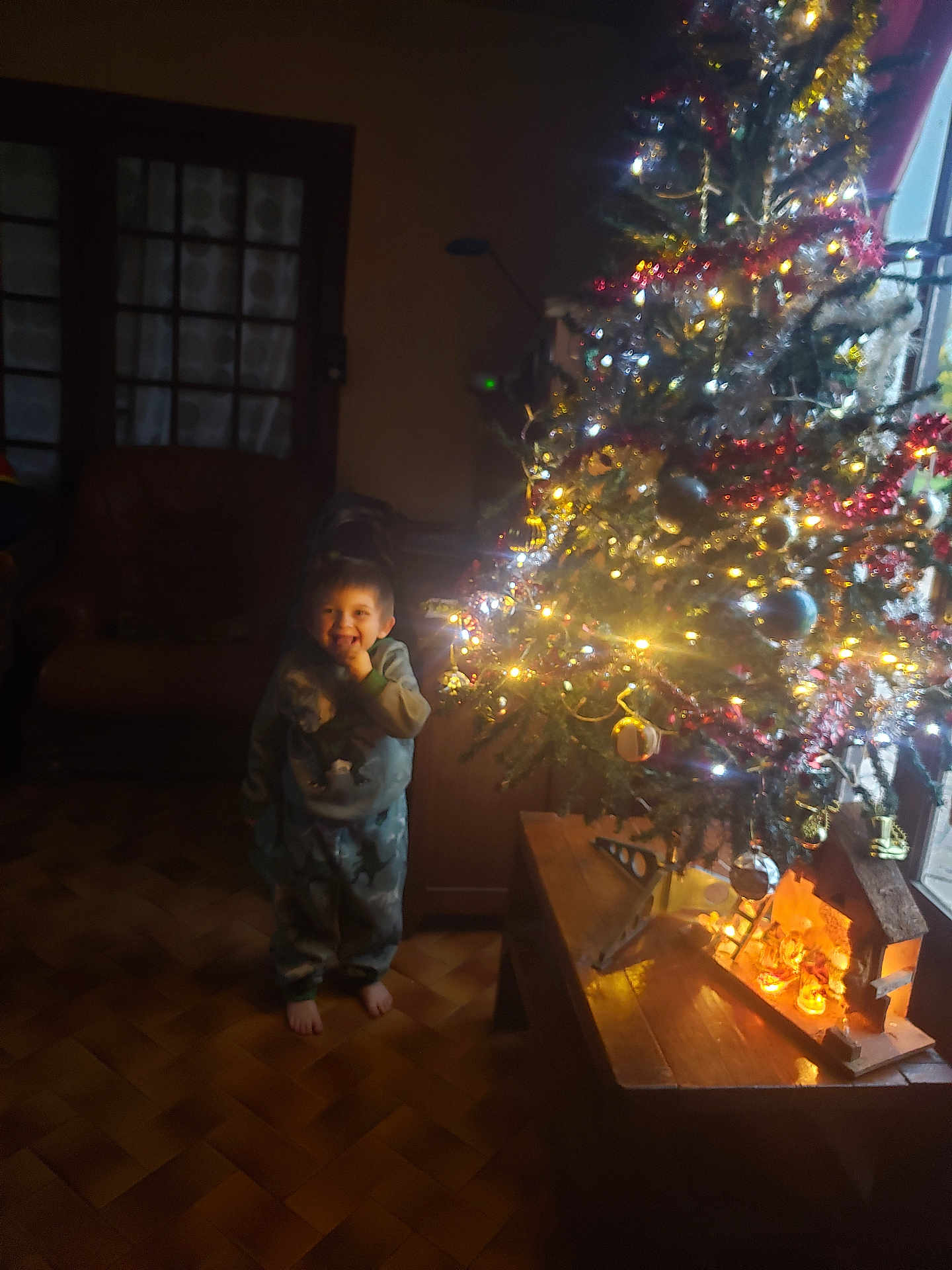 Claude a rejoint le concours — aidez-le/la à gagner de superbes lots ! barefoot, chair, child, christmas_tree, cozy, decorations, festive, furniture, holiday, indoors, lamp, lights, ornaments, pajamas, person, smile, smiling, warm_lighting, window, wooden_floor