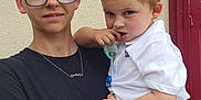 Claude participe au concours pour gagner de l'argent avec cette photo : background, black_shirt, casual, child, clothing, expression, face, glasses, hand, holding, indoor, light_skin, pacifier, person, portrait, red_doorframe, short_hair, teen, toddler, white_polo