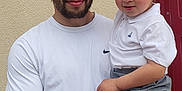 Claude a rejoint le concours — aidez-le/la à gagner de superbes lots ! man, toddler, child, person, holding, white_shirt, polo_shirt, gray_pants, beige_wall, red_door, smiling, curious_expression, outdoor, portrait, people, casual_clothing, short_hair, facial_hair, light_skin, close_up