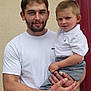 man, toddler, child, person, holding, white_shirt, polo_shirt, gray_pants, beige_wall, red_door, smiling, curious_expression, outdoor, portrait, people, casual_clothing, short_hair, facial_hair, light_skin, close_up