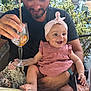 baby, child, man, smile, happy, pink_dress, headband, glass, drink, straw, orange_slice, outdoor, restaurant, table, chair, plant, greenery, person, seated, casual