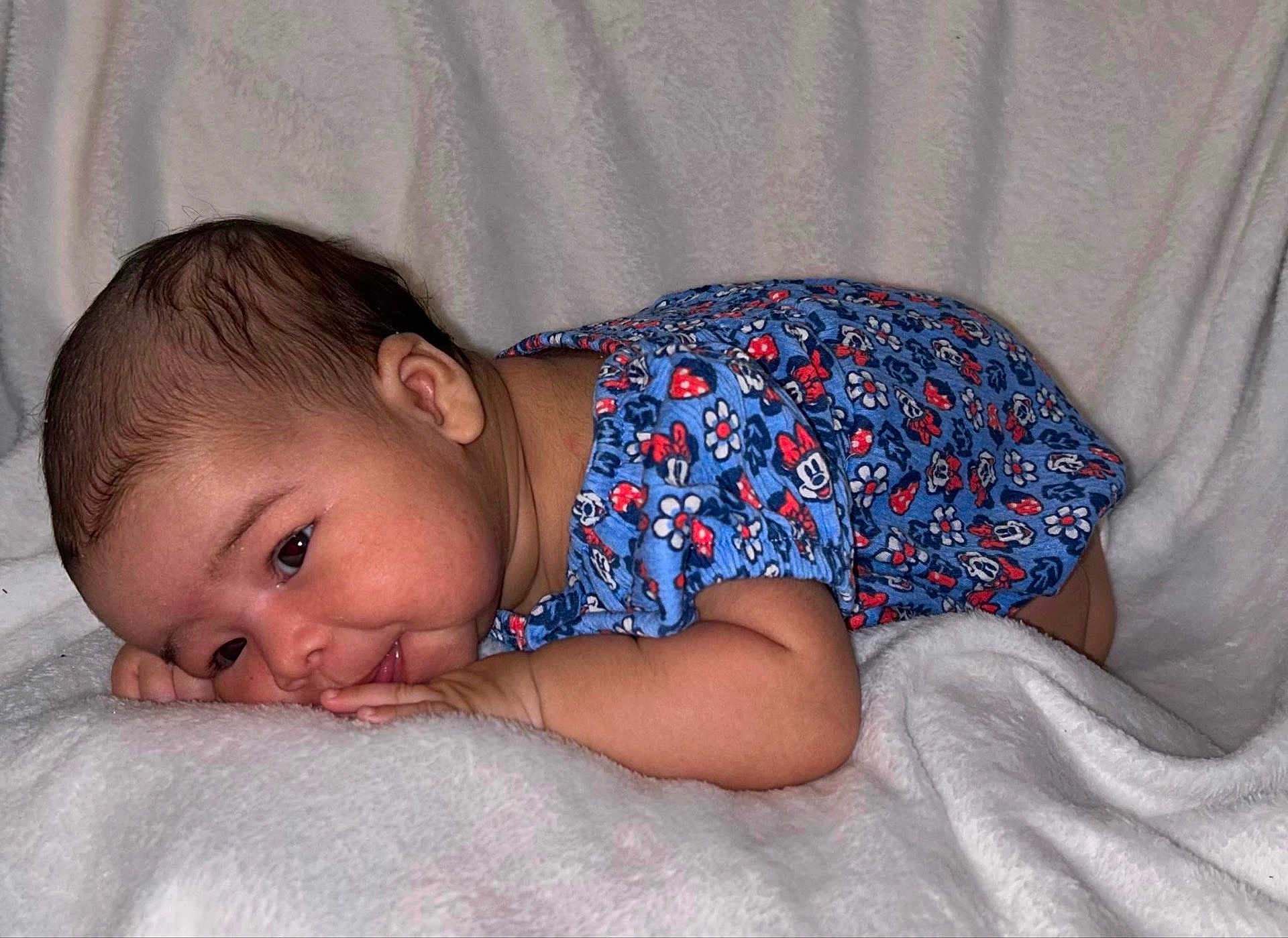 Analaiah joined the competition — help win amazing prizes! baby, child, infant, blue_onesie, floral_pattern, blanket, soft_texture, lying_down, smile, face, head, skin, cute, young, portrait, indoors, cozy, resting, small_hands, expression