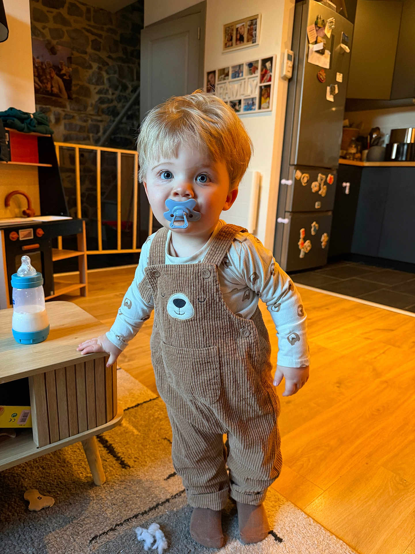 Eden a rejoint le concours — aidez-le/la à gagner de superbes lots ! toddler, child, pacifier, overalls, bear_design, wooden_table, milk_bottle, indoor, kitchen, floor, carpet, fridge, home, toy, socks, baby, cute, standing, curious, light