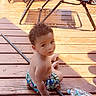 toddler, child, curly_hair, swim_trunks, wooden_deck, outdoor, sunlight, water_bottle, plastic_bottle, metal_rod, folding_chairs, shadow, bare_chest, summer, sitting, looking_up, young_child, casual, daylight, backyard
