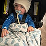 baby, pacifier, hat, blanket, car_seat, blue_pajamas, child, indoor, cute, infant, sitting, warm_clothing, safety_harness, floor, wood_floor, soft_toy, comfort, portrait, expression, hand
