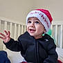 toddler, child, baby, christmas_hat, black_jacket, crib, bed, plaid_pants, wooden_sign, indoor, smiling, happy, cute, person, hand, face, headwear, home, holiday, expression