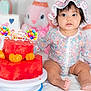 baby, child, birthday_cake, watermelon_cake, fruit, party, hat, floral_clothing, sitting, cute, celebration, indoor, toy, stuffed_animal, colorful, food, table, white_blanket, decorations, happy_birthday