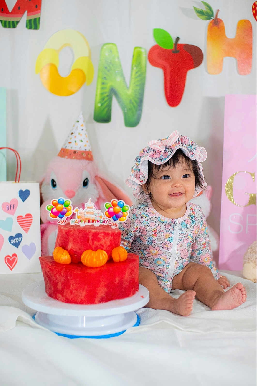 Astoria is registered to the contest to win money with this photo: baby, birthday, cake, watermelon, fruit, party, smiling, child, onesie, bonnet, stuffed_animal, bunny, decorations, colorful, orange, tangerine, celebration, happy, indoor, cute