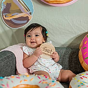 Astoria is registered to the contest to win money with this photo: baby, infant, cute, smiling, hair_clips, wooden_sign, four_months, couch, blanket, donut_decoration, colorful, sitting, indoors, happy, child, portrait, celebration, playful, home, decor