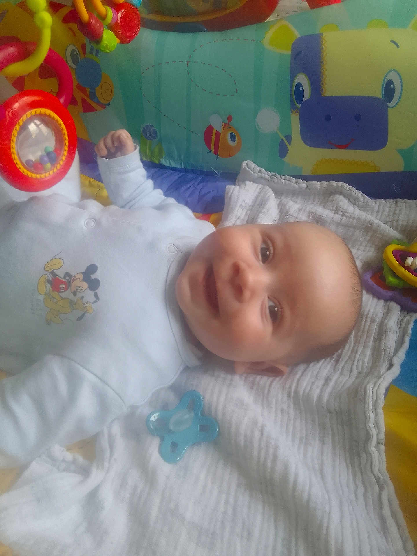Alexander is registered to the contest to win money with this photo: baby, smiling, onesie, mickey_mouse, pacifier, play_mat, toy, colorful, blanket, infant, lying_down, child, face, happy, indoor, cute, head, clothing, soft_toy, baby_toy