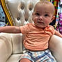 baby, child, toddler, chair, gold, white, plaid_shorts, peach_shirt, furniture, indoor, cute, portrait, sitting, young_child, person, face, expression, curious, comfortable, decor