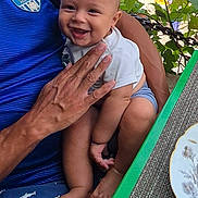 Léandro participe au concours pour gagner de l'argent avec cette photo : baby, child, smiling, person, adult, hand, blue_shirt, shorts, shark_pattern, table, plate, placemat, green_table, outdoor, tree, leaf, stone_wall, scooter, people, sunlight