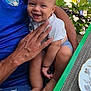 baby, child, smiling, person, adult, hand, blue_shirt, shorts, shark_pattern, table, plate, placemat, green_table, outdoor, tree, leaf, stone_wall, scooter, people, sunlight