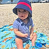 baby, child, beach, sand, towel, hat, blue_shirt, sunny, outdoor, clouds, sky, cute, sitting, animal_print, curious, summer, daylight, person, infant, playful