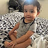 Xavier is registered to the contest to win money with this photo: baby, toddler, child, sitting, bed, blanket, animal_print, curly_hair, smile, indoor, clothing, onesie, person, face, foot, hand, window, natural_light, room, television