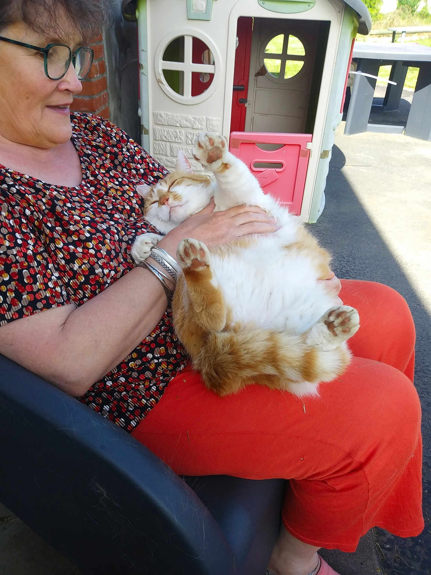 Caramel a rejoint le concours — aidez-le/la à gagner de superbes lots ! woman, cat, glasses, orange_pants, patterned_shirt, bracelets, chair, playhouse, outdoor, sunlight, happy, pet, relaxing, paw, fur, smiling, seated, playful, casual, comfort