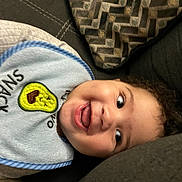 Josiah is registered to the contest to win money with this photo: toddler, child, face, smile, bib, avocado, snack, couch, pillow, curly_hair, happy, indoors, clothing, baby, expression, person, closeup, joy, playful, relaxing