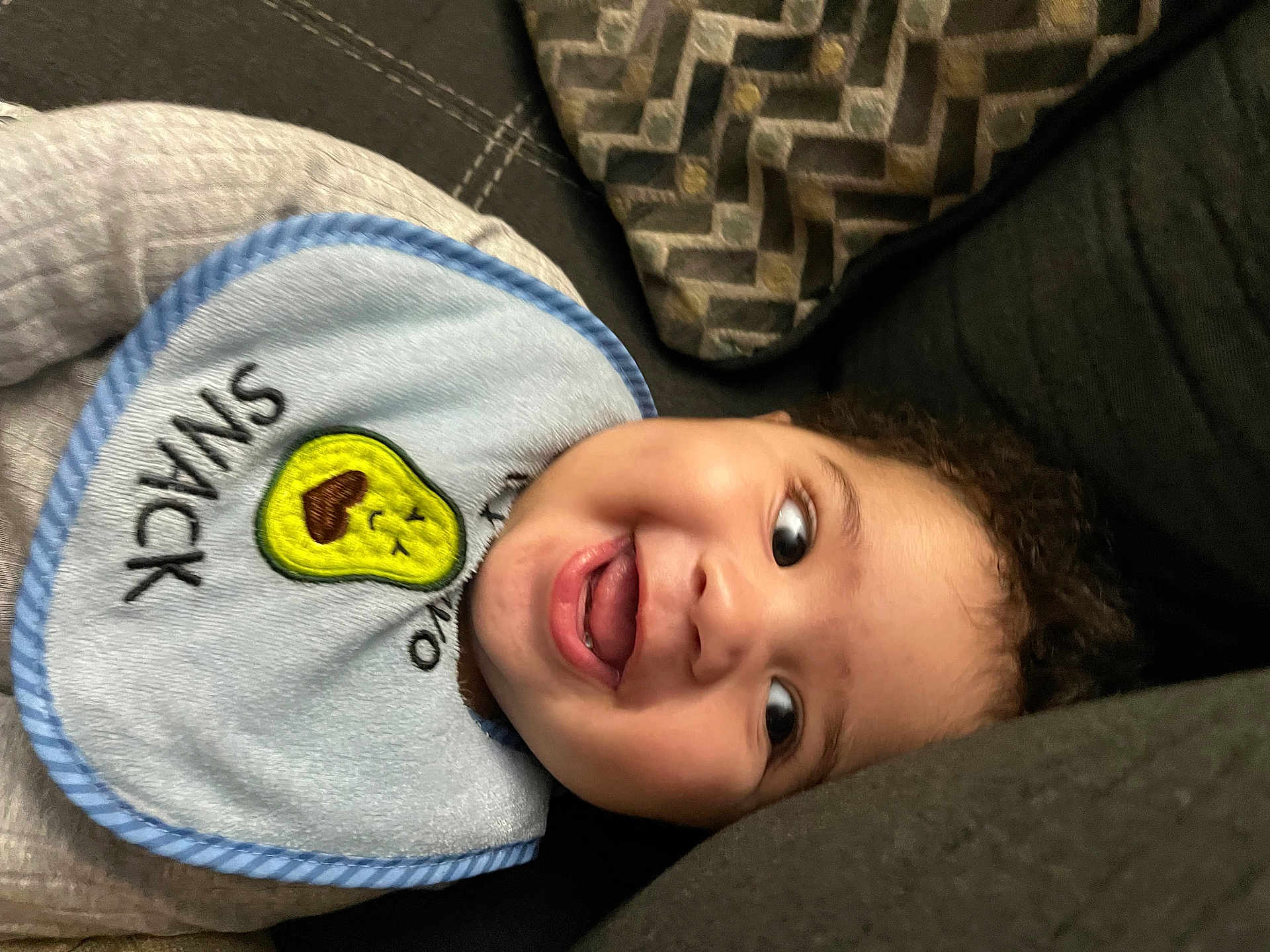Josiah is registered to the contest to win money with this photo: toddler, child, face, smile, bib, avocado, snack, couch, pillow, curly_hair, happy, indoors, clothing, baby, expression, person, closeup, joy, playful, relaxing