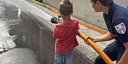 Leo participe au concours pour gagner de l'argent avec cette photo : child, firefighter, fire_hose, water, wet_pavement, concrete_wall, outdoor, person, shorts, red_shirt, footwear, teamwork, learning, spraying, hose, activity, summer, safety, fun, training