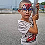 child, boy, face_paint, glasses, tshirt, jeans, pole, outdoor, parking_lot, asphalt, car, fence, building, sky, short_hair, casual_clothing, portrait, holding, curious, daylight