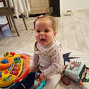 Alice participe au concours pour gagner de l'argent avec cette photo : baby, child, indoor, toy, blanket, floor, play, happy, tongue_out, clothing, person, cute, smile, sitting, wood_floor, furniture, colorful, soft_toy, rattle, home