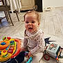 baby, child, indoor, toy, blanket, floor, play, happy, tongue_out, clothing, person, cute, smile, sitting, wood_floor, furniture, colorful, soft_toy, rattle, home