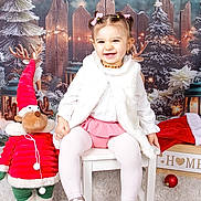 Matilde a rejoint le concours — aidez-le/la à gagner de superbes lots ! toddler, child, smile, white_vest, pink_skirt, pink_bows, chair, plush_reindeer, red_clothing, green_clothing, christmas_decor, holiday, winter, snow, lights, carpet, ornament, background, festive, indoor