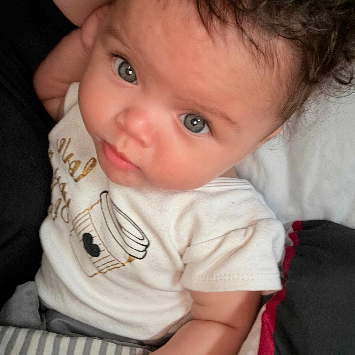 Arie joined the competition — help win amazing prizes! baby, child, infant, face, eyes, curly_hair, white_shirt, coffee_cup_design, blanket, striped_blanket, bed, cute, portrait, indoor, skin, person, young_child, expression, looking_up, cozy