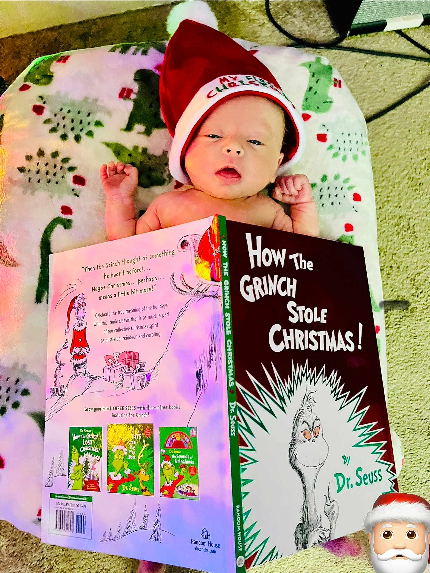 Anakin joined the competition — help win amazing prizes! baby, infant, hat, santa_hat, book, how_the_grinch_stole_christmas, dr_seuss, blanket, dinosaur_pattern, carpet, reading, holiday, christmas, child, cute, indoors, person, face, lying_down, cozy