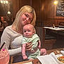 baby, child, woman, restaurant, table, food, drink, wooden_wall, chandelier, menu, napkin, plate, bracelet, blonde_hair, casual_clothing, indoor, people, seated, holding, portrait