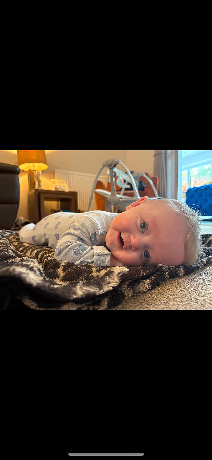 George is registered to the contest to win money with this photo: baby, blanket, indoor, living_room, furniture, lamp, baby_swing, carpet, smiling, face, child, person, blue_eyes, onesie, curly_hair, head, lying_down, soft_toys, cozy, natural_light