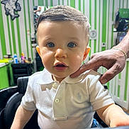 Jesus is registered to the contest to win money with this photo: toddler, child, boy, white_polo_shirt, haircut, barber_chair, adult_hand, blue_eyes, short_hair, indoor, green_wall, striped_wall, mirror, salon, person, portrait, curious, candid, clothing, furniture